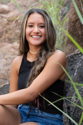 A person with long hair sitting outdoors, wearing a black sleeveless top and denim shorts, surrounded by rocks and grass.