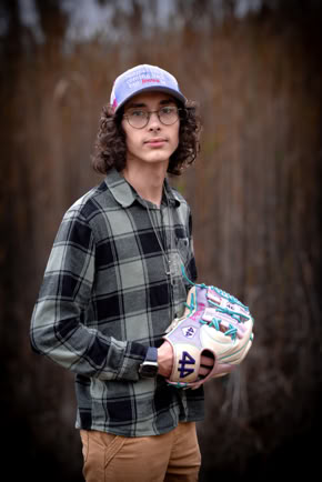 A person with long curly hair wearing glasses, a plaid shirt, and a cap, holding a colorful baseball glove, standing outdoors.