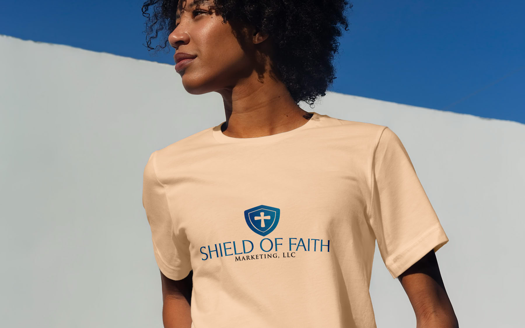 Person with curly hair wearing a beige t-shirt with a shield logo and text, standing against a blue and white background.
