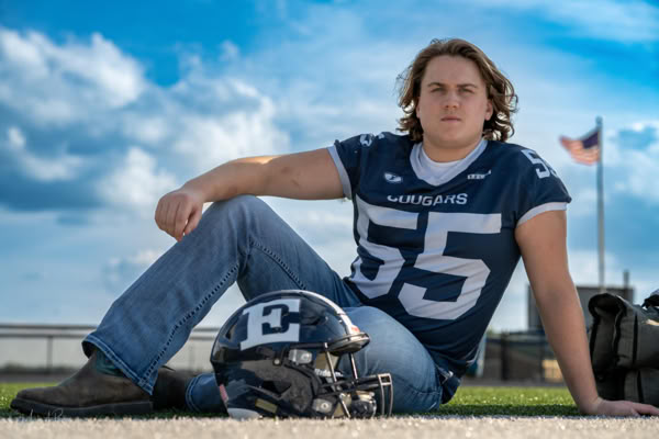 A person wearing a football jersey with the number 55 sits on the ground next to a helmet, with a cloudy sky and an American flag in the background.