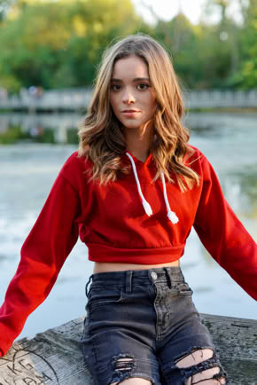 Young person with long hair wearing a red hoodie and ripped jeans, sitting outdoors near a body of water with trees in the background.