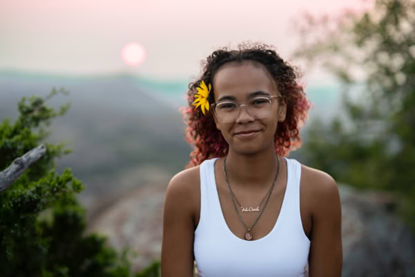 A person with curly hair and a yellow flower tucked behind the ear, wearing glasses and a white tank top, stands outdoors with a blurred natural landscape and sunset in the background.
